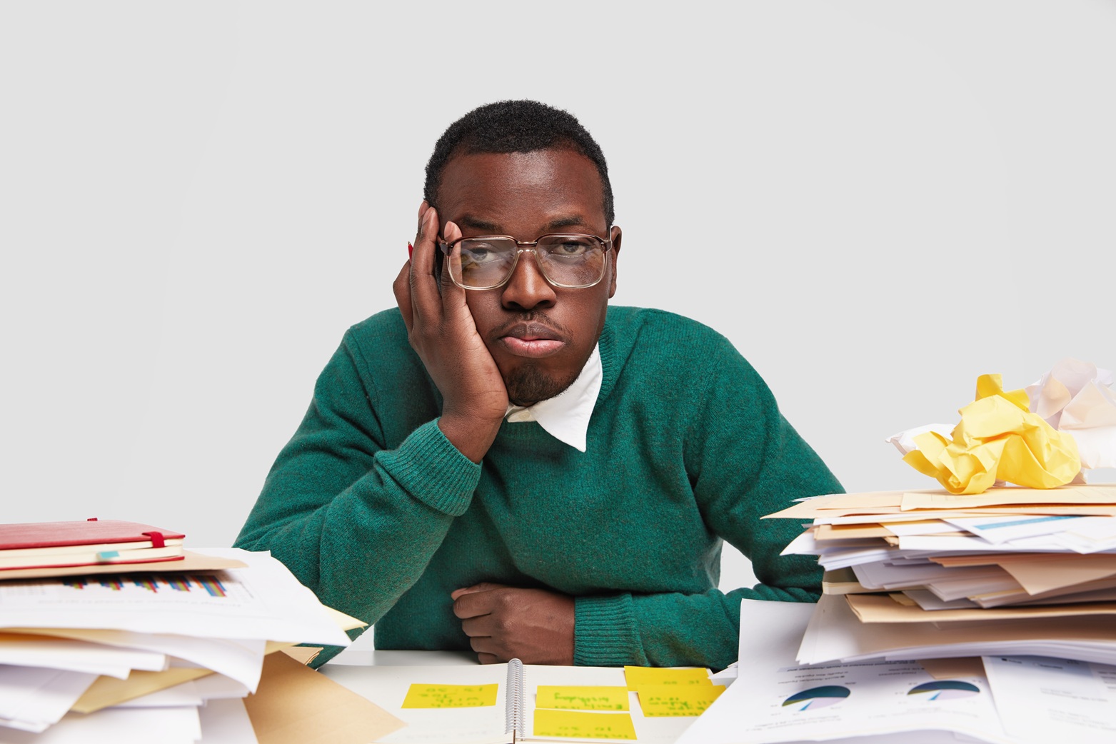 Bored male worker has tired facial expression, looks in displeasure, works long hours on project task, has dark healthy skin, sticks notes in notepad, tries not to forget everything, isolated